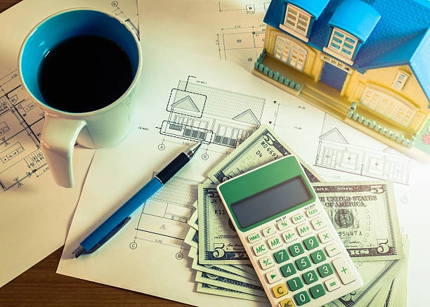 Arhitekturni načrt, denar, kalkulator, maketa hiše in skodelica kave na mizi arhitekta.