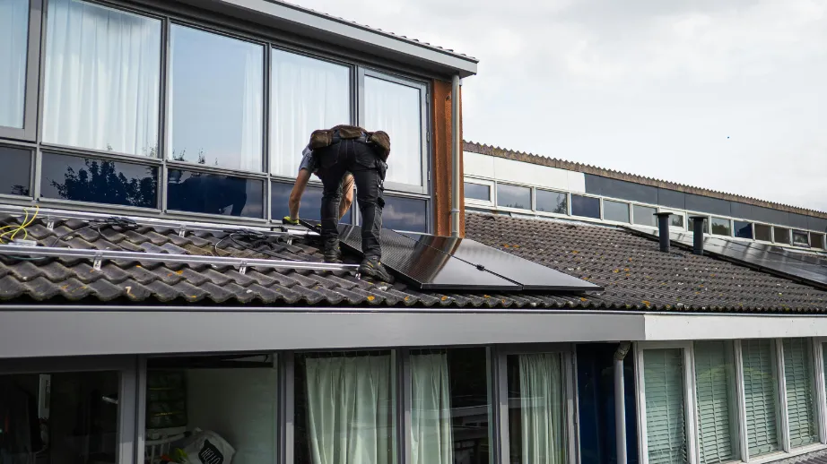 Monter na strehi iz strešnikov namešča sončni panel na pritrdilne nosilce ob nadstropnih oknih; vidni so kabli in delno nameščeni moduli.