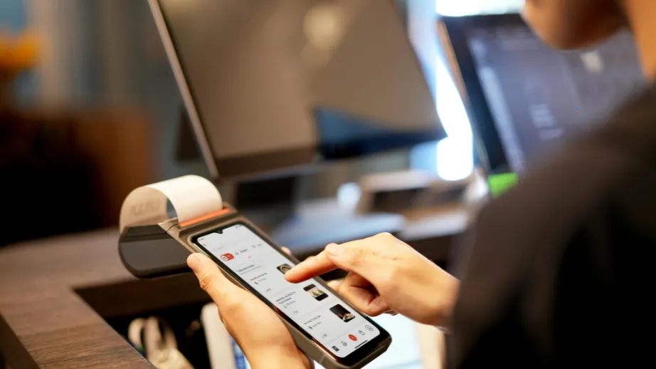 Oseba pri blagajni uporablja mobilni telefon za plačilo, ob blagajniškem terminalu in računalniškem zaslonu, kar ponazarja sodobno brezstično plačevanje.