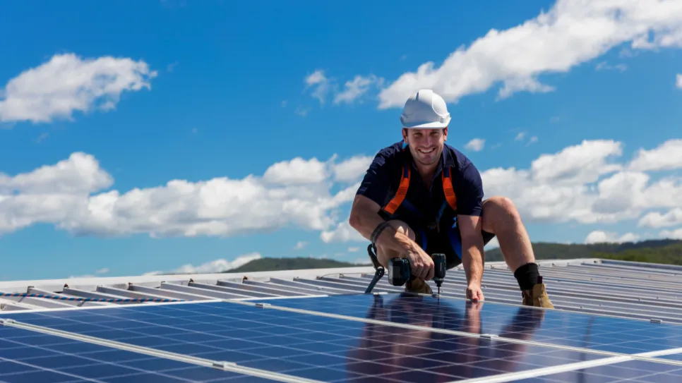 Monter pritrjuje sončne kolektorje na streho. Gleda v kamero in se smeji.