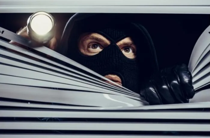 Vlomilec z žepno svetilko in kapuco na glavi gleda skozi rolete na oknu.