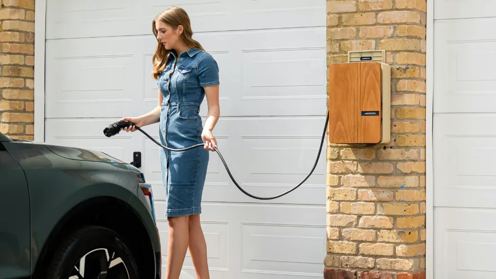 Ženska priklopi električni avtomobil na domačo električno polnilnico. Domače polnilnice omogočajo priročno in cenovno ugodno polnjenje električnih vozil.