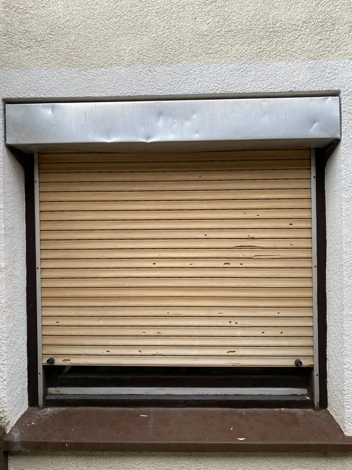 A closed window shutter with visible dents and wear on a building exterior.