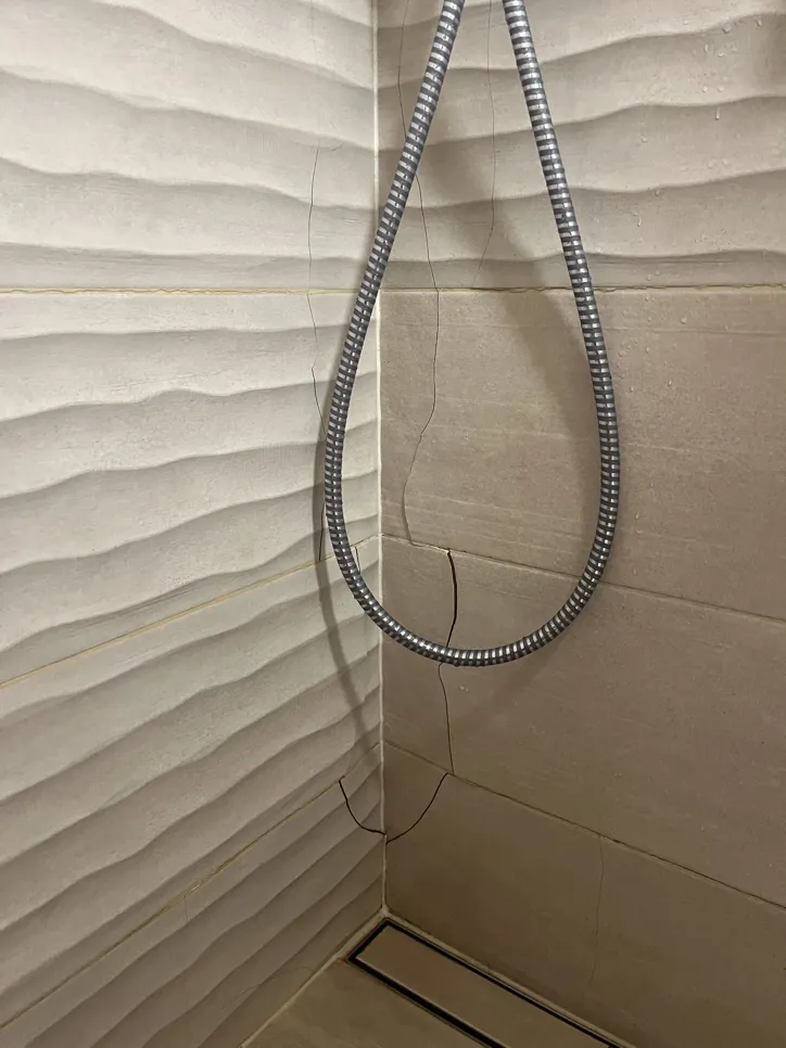 Cracked tiles in a shower corner with a coiled metal hose hanging nearby, indicating potential water damage.