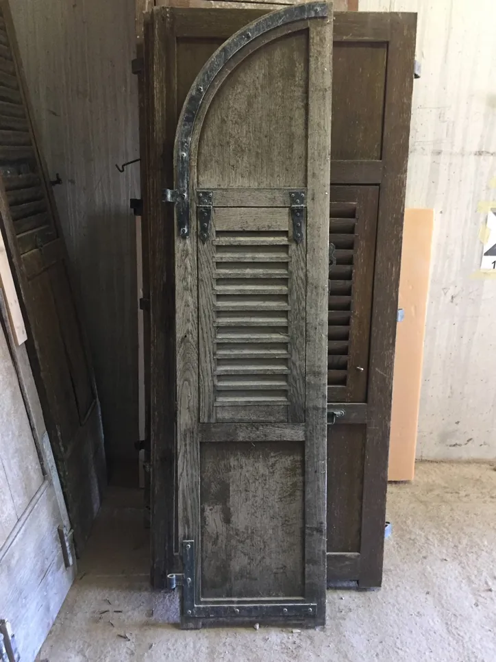 Old wooden shutter with arched top and weathered finish, featuring metal hinges and slats.