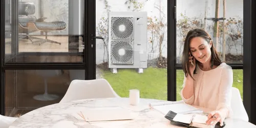 Ženska v notranjosti govori po telefonu, čez stekleno okno se vidi zunanja enota toplotne črpalke.
