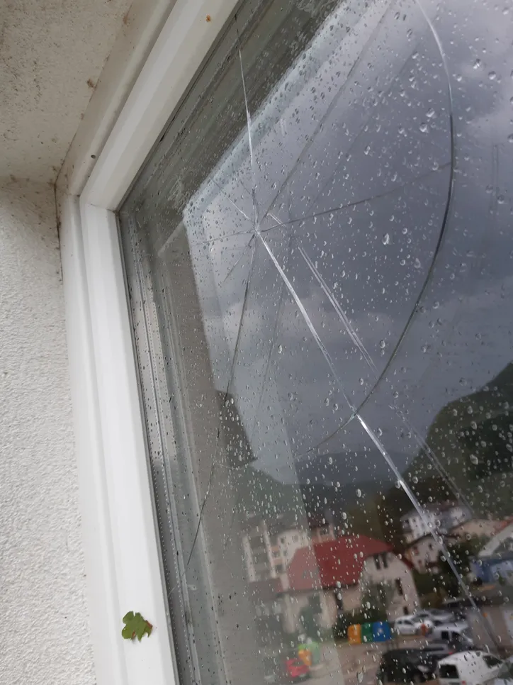 A close-up of a cracked window with rain droplets, set against a stormy sky and buildings in the background.