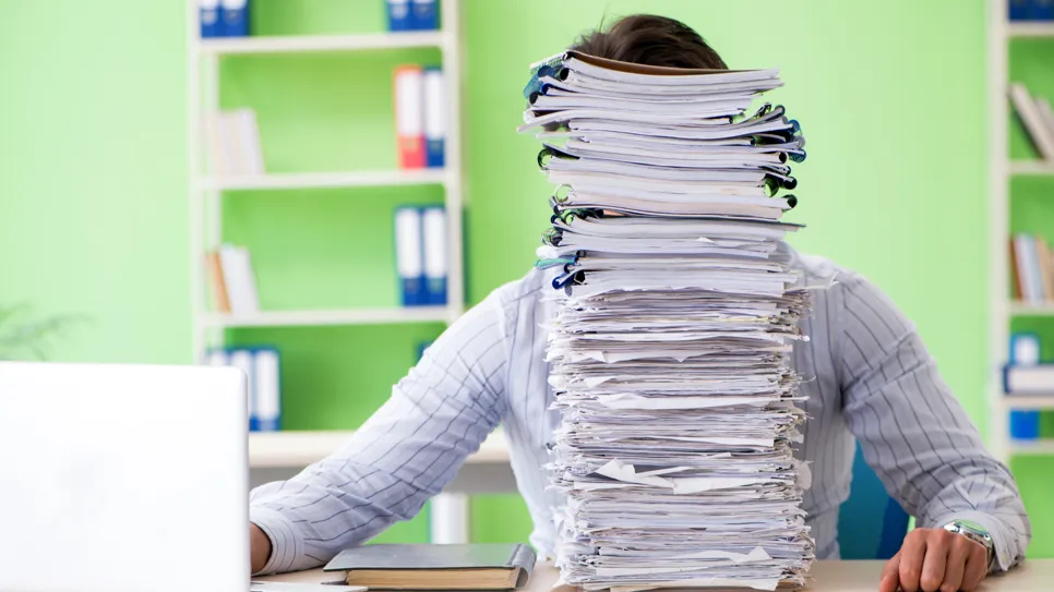 moški za pisalno mizo, ki ga zakrivajo visoko naloženi papirnati dokumenti