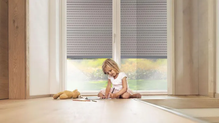 Punčka se igra na tleh v notranjosti sobe, ki je zasenčena z zunanjimi roletami.