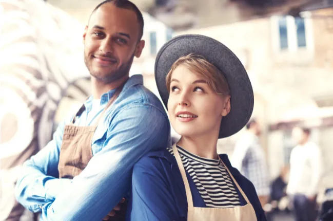 Mlada fant in dekle v predpasnikih stojita z ramo ob rami v svojem lokalu.