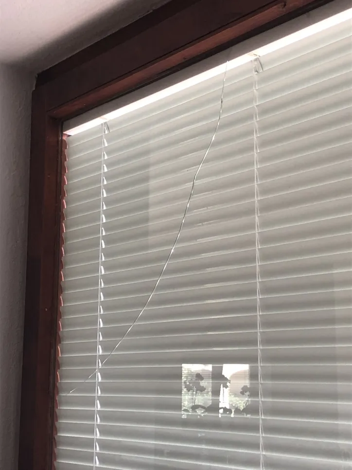 A window pane with a visible crack runs vertically behind white blinds in wooden frame.