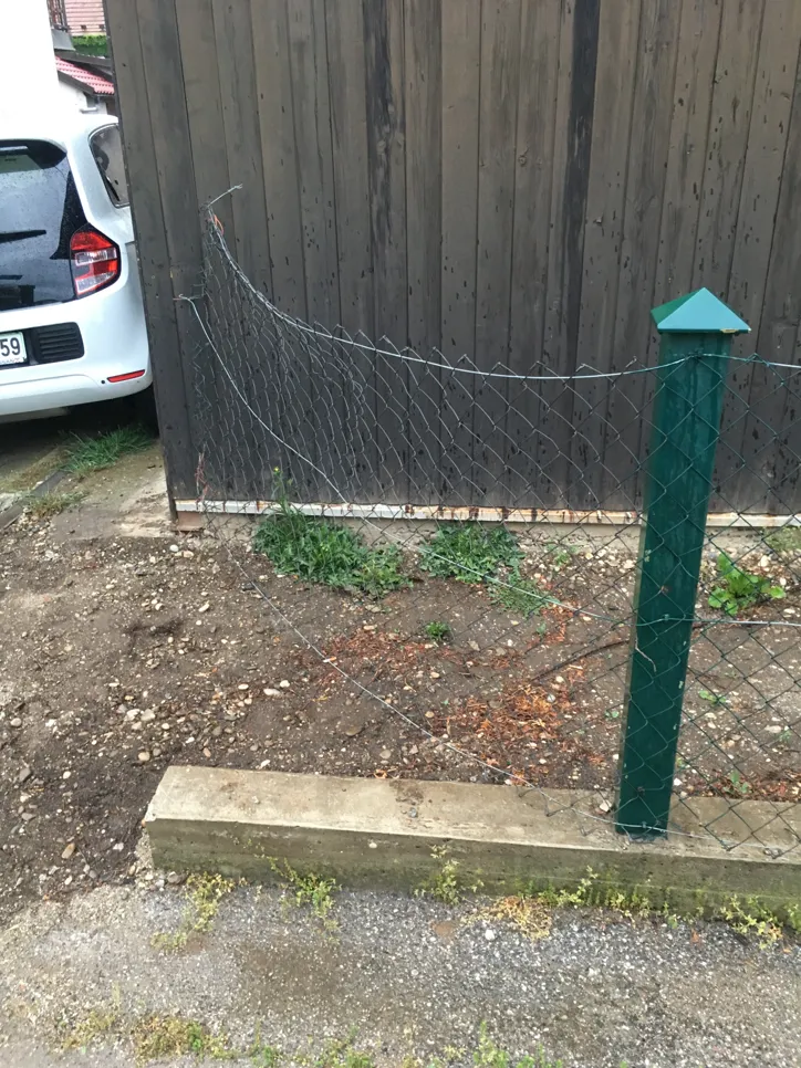 Bent chain link fence next to a parked white car and wooden structure, indicating possible collision damage.
