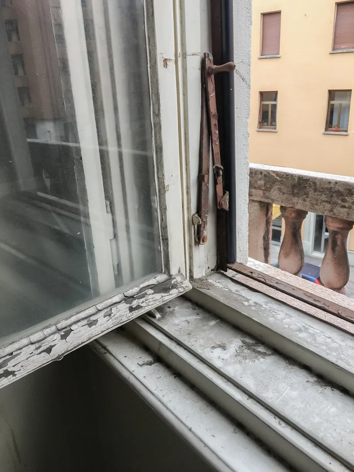 Old window with peeling paint and rusty hinge, indicating deterioration and potential draft issues.