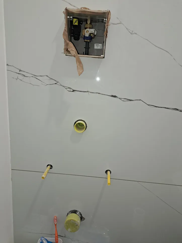 Nedokončana namestitev vodovodne napeljave v kopalnici z razkritimi cevmi in električnimi napeljavami na ploščicah.