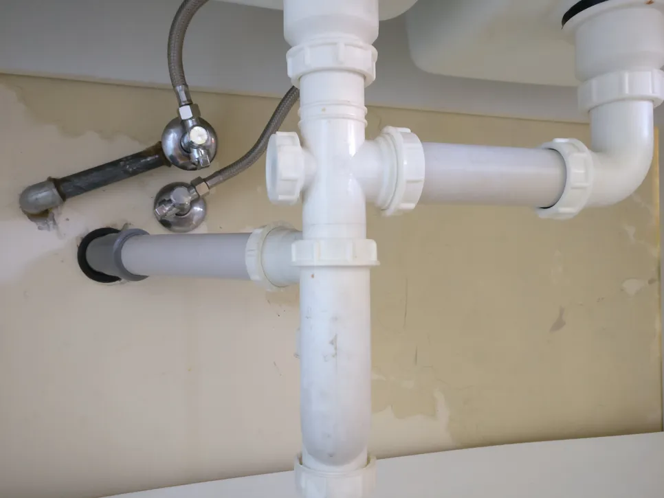 Pogled od blizu na cevi pod umivalnikom z vidnimi cevmi in priključki, ki kažejo na možnost puščanja.